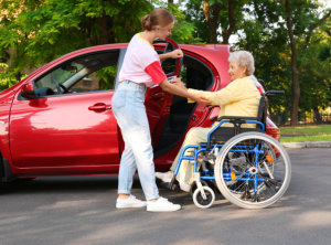 Young women helping grandmother in wheelchair to get into car outdoors