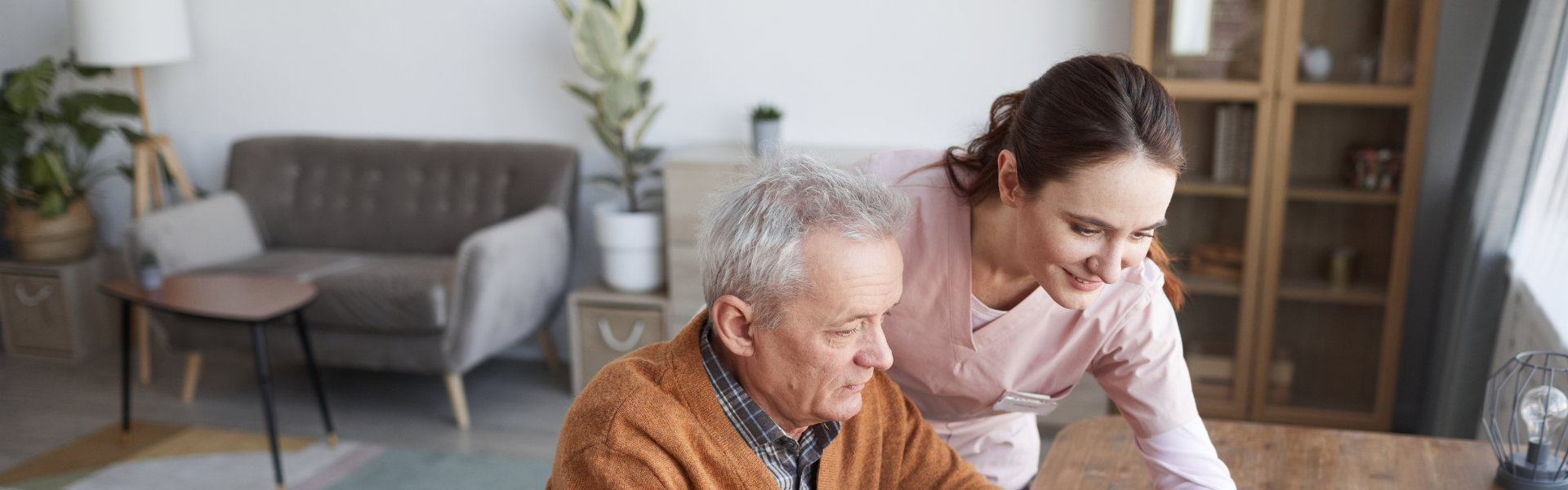 Portrait of senior man in wheelchair using laptop at retirement home with nurse assisting him