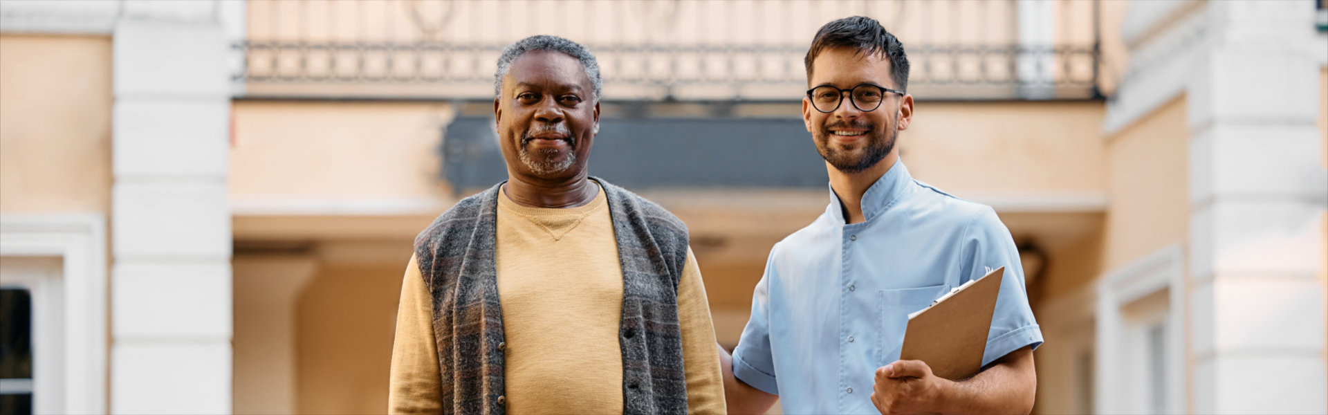 senior man and young caregiver in front of residential care home looking at camera